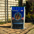 A large bag of Nutrena Respond Original Beef-Pulp Feed stands on a stone patio in front of a fence and garden plants.