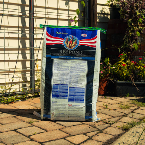 A large bag of Nutrena Respond Original Beef-Pulp Feed stands on a stone patio in front of a fence and garden plants.