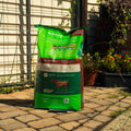 A large bag of Natural Balance Safe-Choice All Life Stages Premium Horse Feed stands on a stone patio in a backyard setting with a fence and plants in the background.