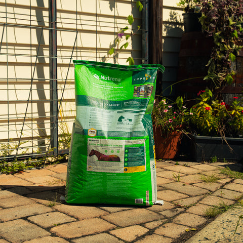 A large bag of Empower Topline Balance supplement stands on a paved patio in front of a garden fence and trellis, with plants visible in the background.