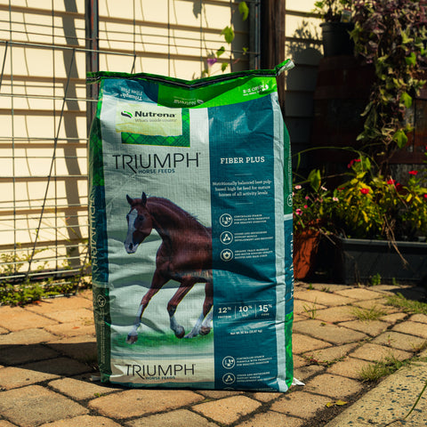 A large bag of Triumph Fiber Plus supplement stands on a paved patio in front of a garden fence and trellis, with plants visible in the background.