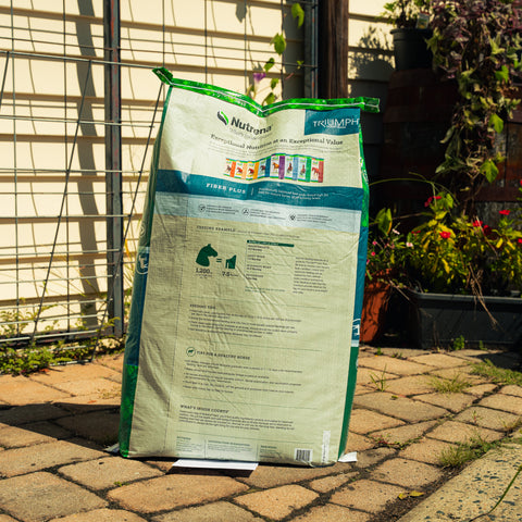 A large bag of Triumph Fiber Plus supplement stands on a paved patio in front of a garden fence and trellis, with plants visible in the background.