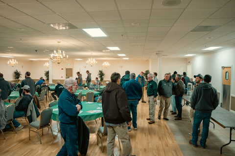Group of people in a large room with tables and chairs, possibly in a community center or similar setting.