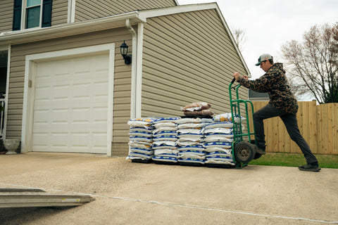 Person pushing a dolly with bags of fertilizer in front of a house.
