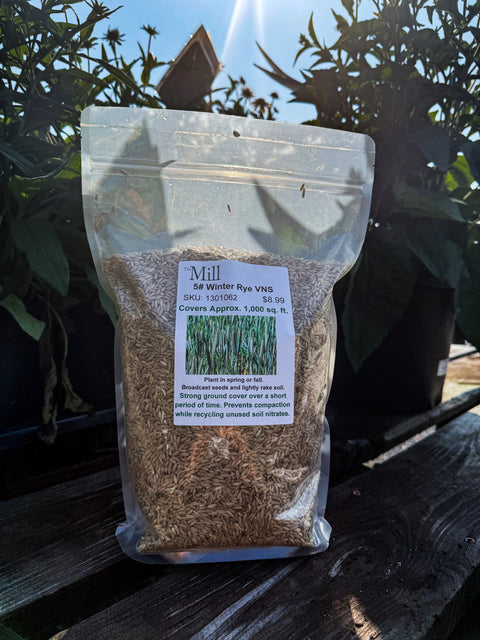 A clear stand-up pouch filled with winter rye sits on a wooden deck outdoors, with potted plants and blue sky visible in the background. The front label reads Mill and shows a grass graphic.
