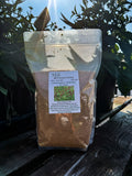A clear stand-up pouch containing brown Mill Clover seed, labeled 'Mill Clover', placed on a dark wooden deck with green plants in the background.