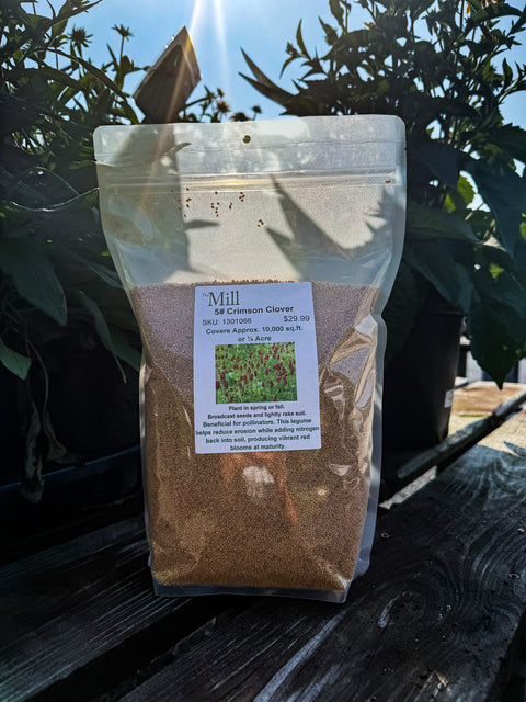 A clear stand-up pouch containing brown Mill Clover seed, labeled 'Mill Clover', placed on a dark wooden deck with green plants in the background.