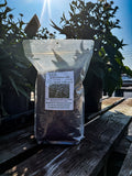A clear stand-up pouch filled with dark brown buckwheat sits on a weathered wooden table outdoors, with potted plants and bright sunlight in the background.