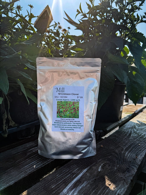 A silver stand-up pouch labeled 'Mill Crimson Clover' sits on a dark wooden bench outdoors, with potted plants and bright sunlight in the background.