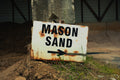 Rust-covered sign pointing to 'Mason Sand' with a arrow on a dirt ground.