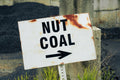 A weathered white metal sign with rusted edges, reading 'NUT COAL' in bold black letters above a black arrow pointing right, mounted on a post outdoors near piles of dark coal.
