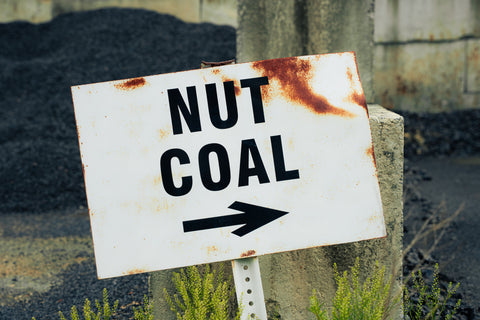 A weathered white metal sign with rusted edges, reading 'NUT COAL' in bold black letters above a black arrow pointing right, mounted on a post outdoors near piles of dark coal.