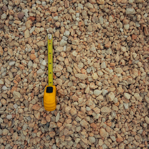 A yellow tape measure extended vertically through a dense bed of small tan, beige, and light brown gravel stones.