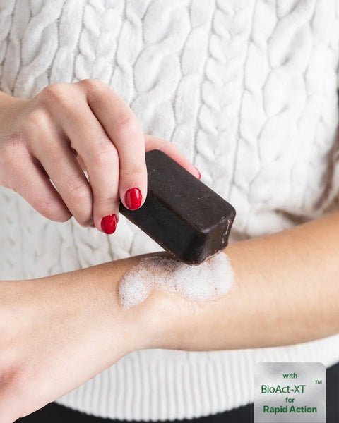A close-up of a person applying a dark brown rectangular foam product onto their forearm, which is covered in white foam. The person is wearing a white cable-knit sweater and has red nail polish.