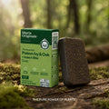 Marie Originals Poison Ivy & Oak + Insect Bite Soap shown with its green box and a dark brown rectangular soap bar placed on a mossy log in an outdoor forest setting.