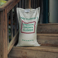 A 50 lb bag of Oregon Grown Clover (Red Clover) standing on rustic wooden steps next to a wooden railing.