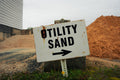 Rustic 'Utility Sand' sign with an arrow pointing to the right, set against a backdrop of sand and concrete.