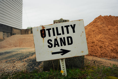 Rustic 'Utility Sand' sign with an arrow pointing to the right, set against a backdrop of sand and concrete.